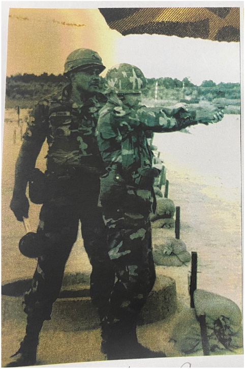 Old photo of two soldiers. One, in side profile, holds a gun with both hands, aiming forward. The other stands behind, gesturing and instructing other soldier.