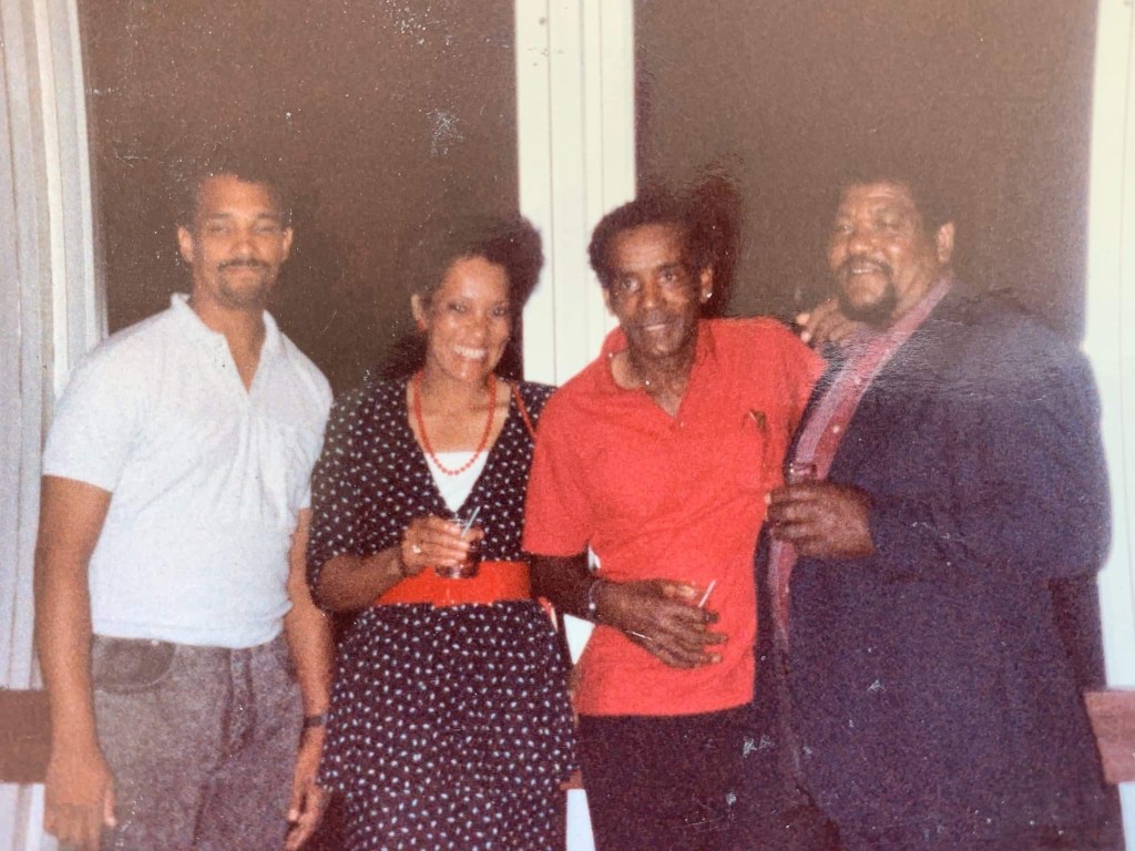Image of four people, three men and one woman smiling into camera in front of window. 