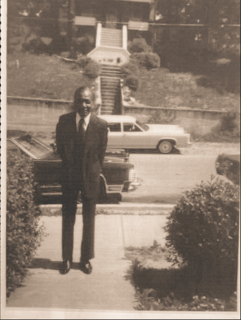 Man wearing a suit posed with his hands behind his back standing on a walkway in front of a neighborhood street lined by two cars