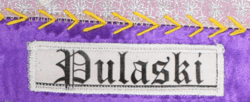 Black text on white background spelling Pulaski. Stitched onto purple quilt