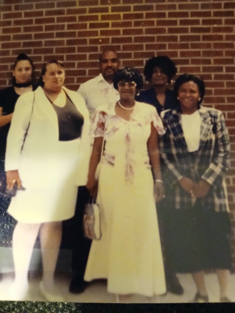 older photo of six people posing for a photo in front of a brick wall