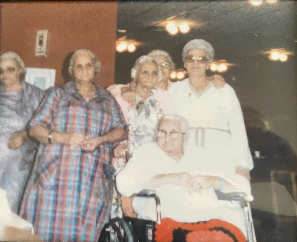 Candid photo depicting six older women wearing dresses. One woman is seated in a wheelchair in front of the other women. Each woman is gazing in different directions.