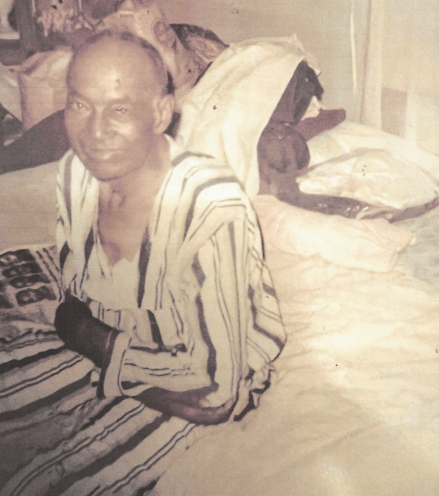 Older man sitting on a the edge of a bed with his arms crossed dressed in pajamas with clutter in the background