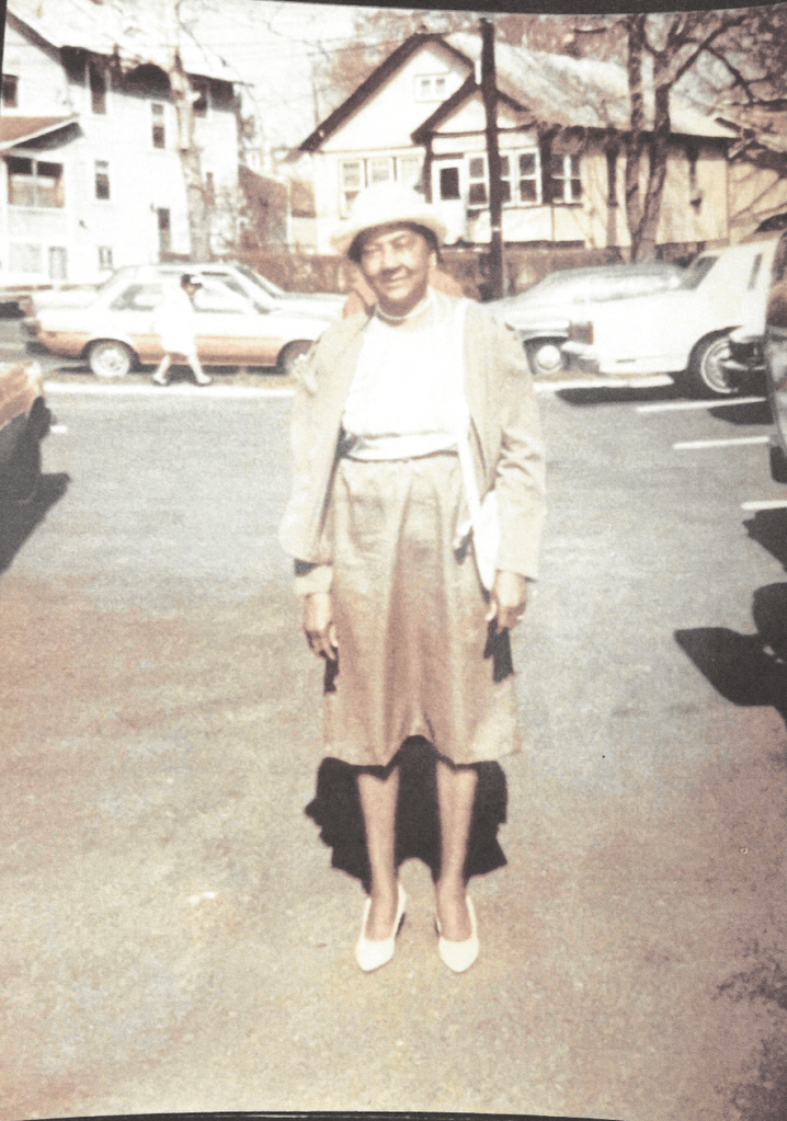 Old photo of a woman smiling and standing in a parking lot with cars and houses in the distance she is dressed in a hat, blouse, skirt, overcoat, and heels.