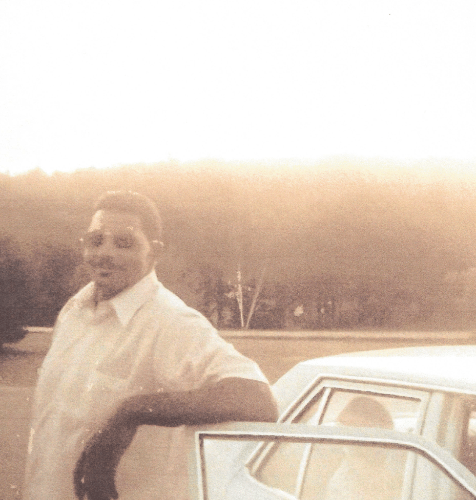 Man standing outside leaning on the passenger door of a car wearing sunglasses in a white shirt