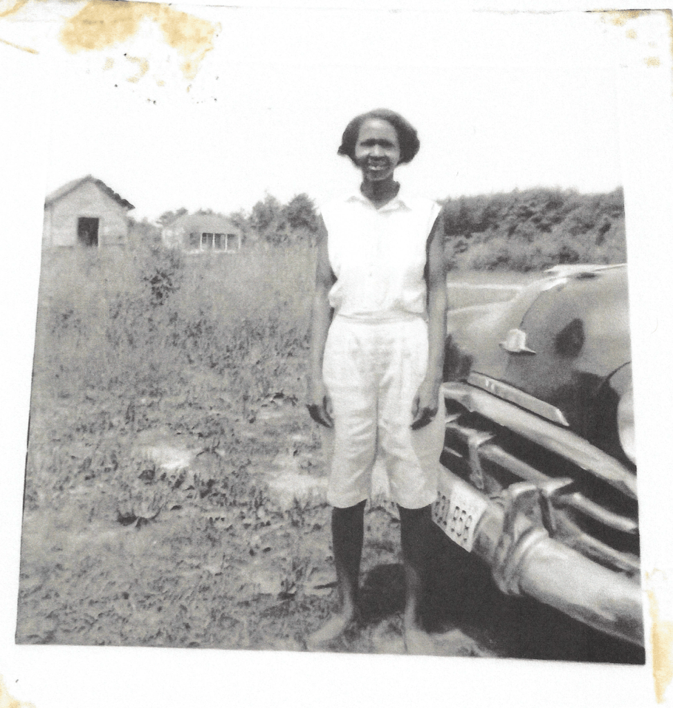 Old black and white photo of a woman dressed in white clothing standing in a field with a barn in the distance and the front bumper of a vehicle adjacent to her. She is standing with her arms by her side, smiling and squinting.