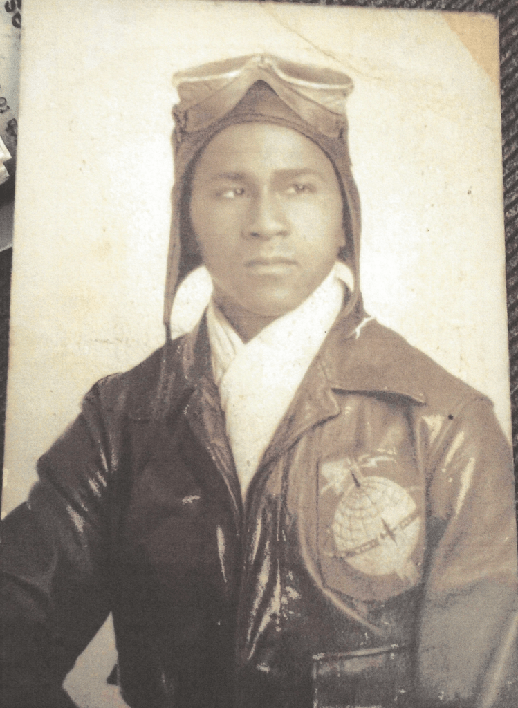 Old black and white photograph of a young man dressed in an aviator uniform with a leather jacket, a hat, and goggles sitting for a portrait