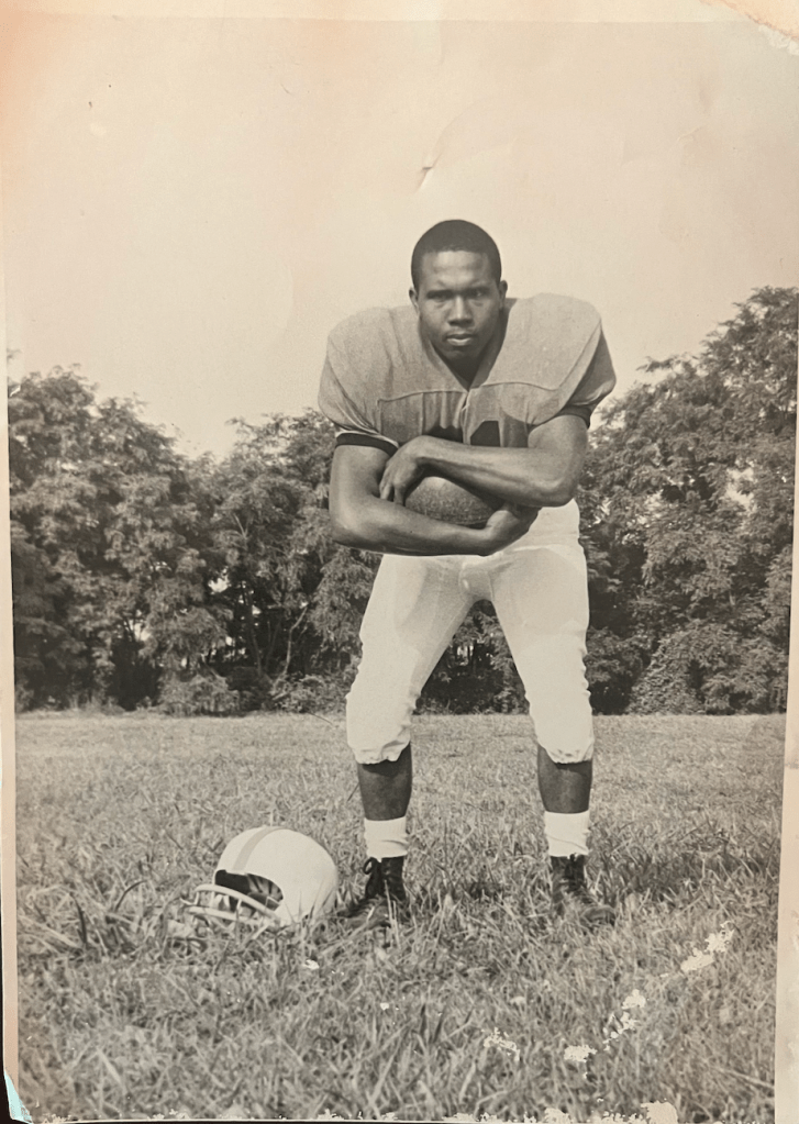 Old black and white photo of young man in football uniform posing outdoors. Hunched over clutching a football in his arms with a helmet lying on the ground beside him. 