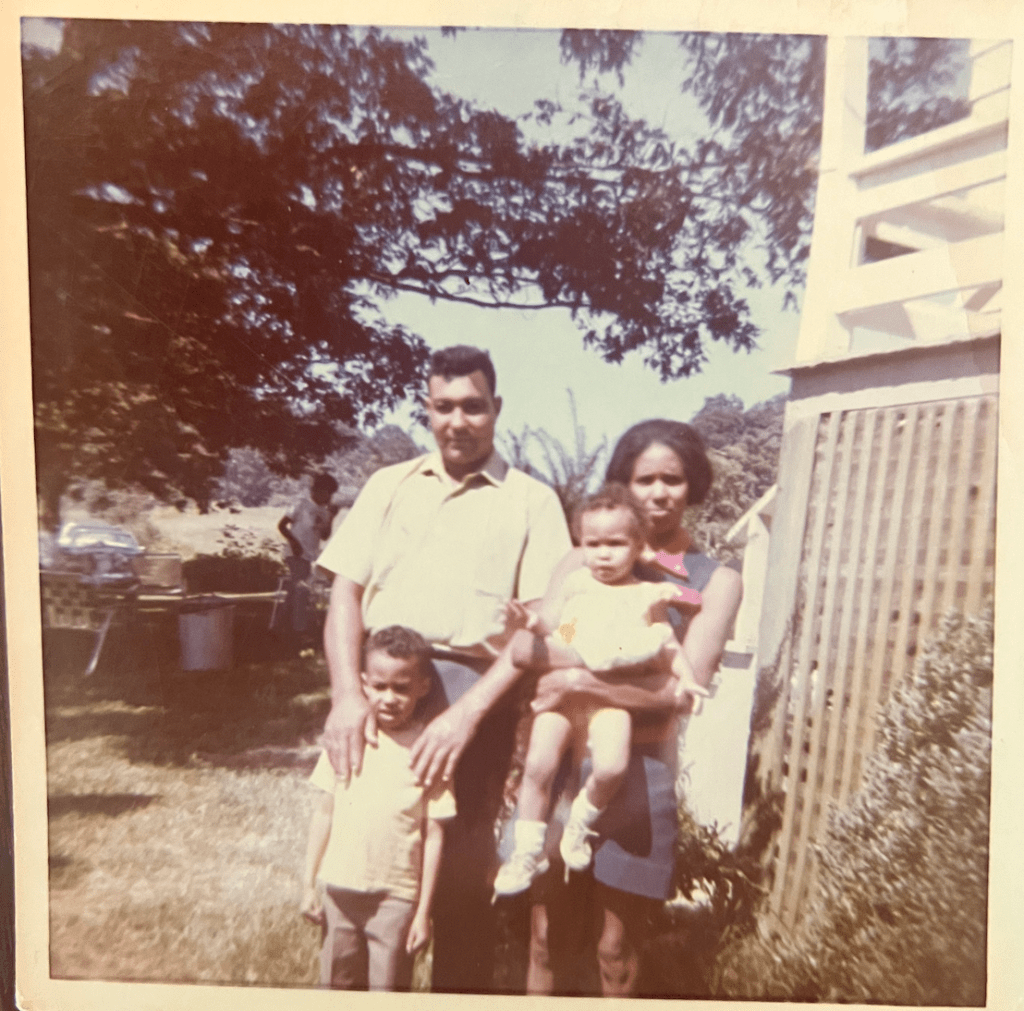 Two adults, a man and a woman standing outside. The man has is hands on the shoulders of a young child below him, while the woman is holding another child in her arms.