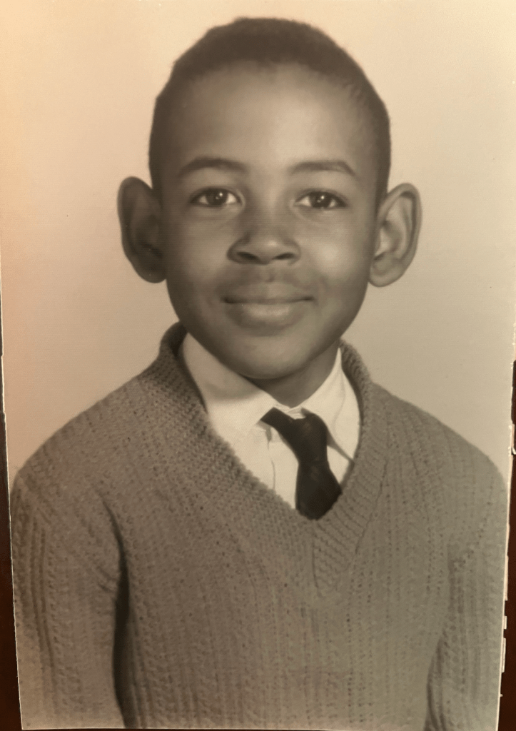Black and white photo of young boy sitting and wearing a sweater over a dress shirt and tie smiling for a portrait.