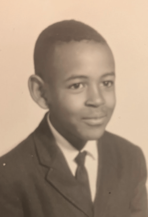 Black and white photo of young boy wearing a suit jacket and a tie sitting and ginning for a portrait looking off to the right.