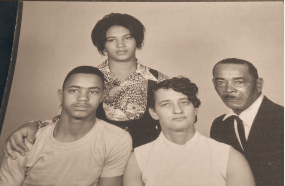 Black and white family portrait. Seated in the front row are a young man, his mother, and his father. Standing in the second row is a young woman with her hand on her brother's shoulder. 