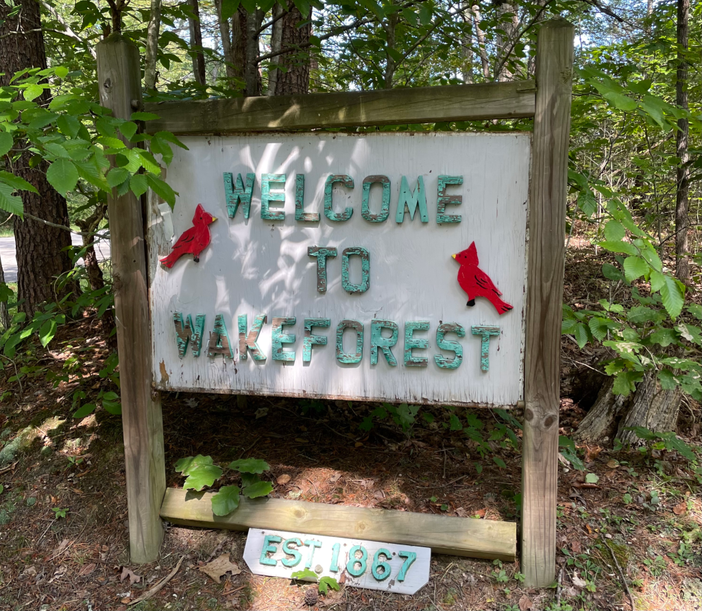 Wooden sign in forest with 'Welcome to Wake Forest' in green and two cardinals on each side