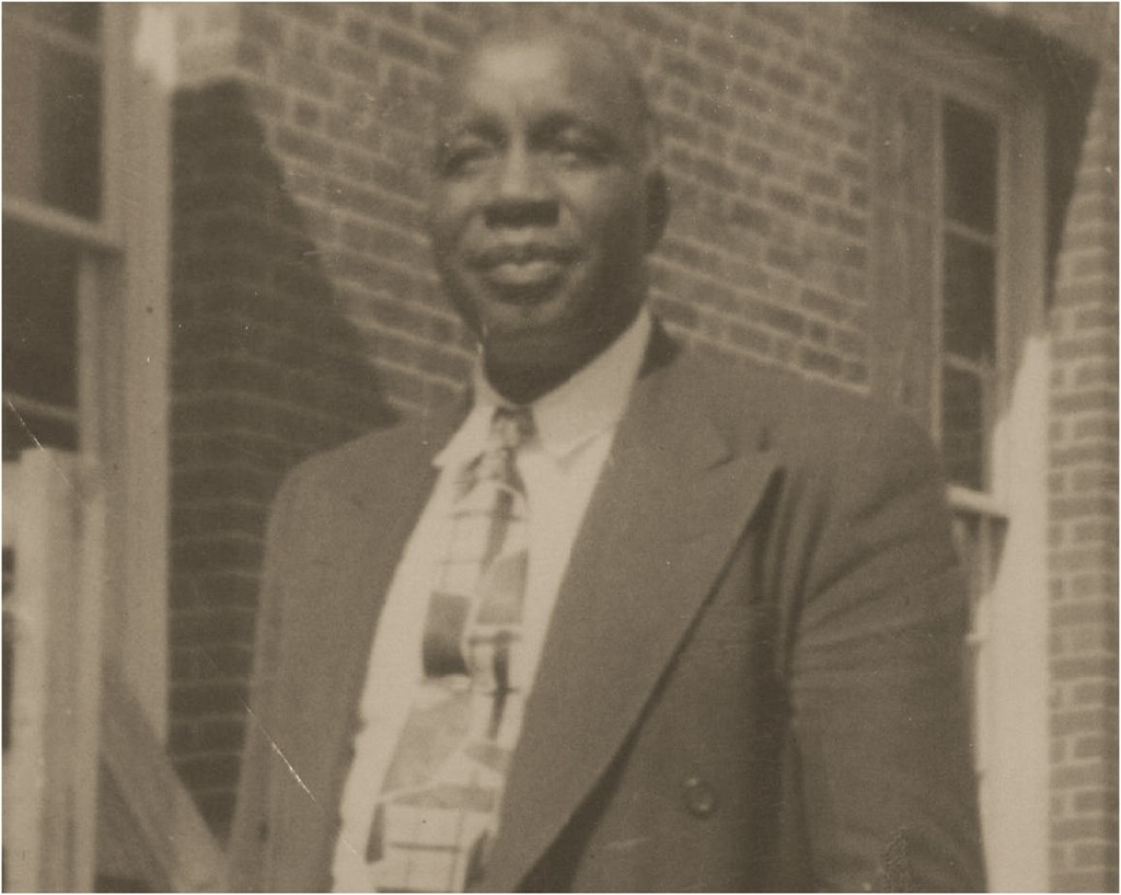 Image of an older gentleman standing in front of a brick building wearing a suit and patterned tie gazing into the distance grinning. 