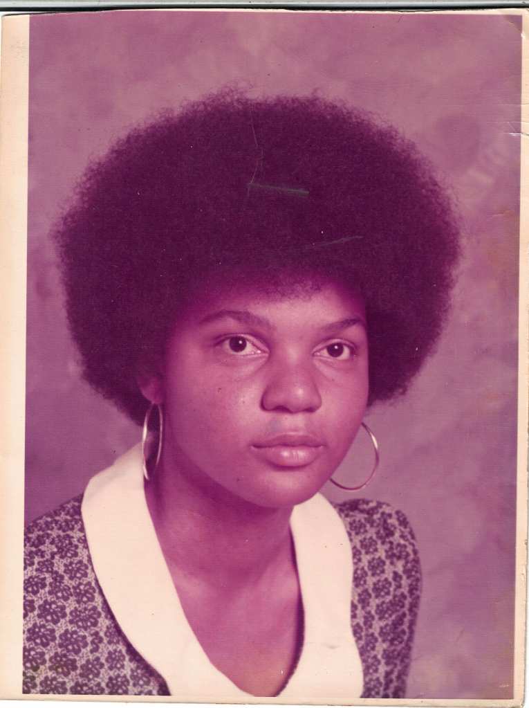 Young Black woman with an afro and hoop earrings, in a patterned dress with round collar, gazing to the right.