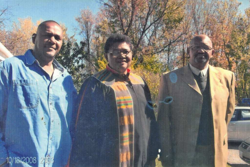 Three people stand side by side outdoors. The men wear a denim shirt and a beige suit jacket while the woman in the middle wears a black robe with African colors.