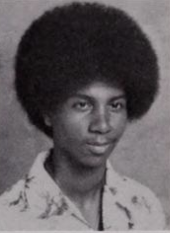Black and white yearbook photo of a young man with an afro wearing a patterned collared shirt grinning 