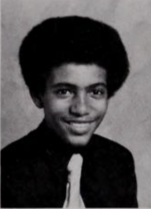Black and white yearbook photo of a young man with dark eyebrows and an afro wearing a black dress shirt and light-colored tie smiling