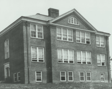 Black and white photo of a three-story building with many windows, a triangular gable, and a chimney.