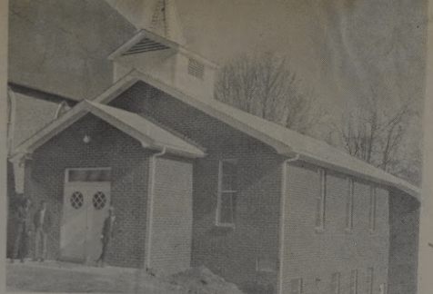 Old photo of baptist church with three people standing in front of front doors with round windows