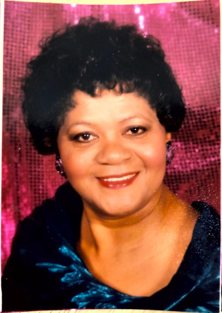 Image of African American woman with curly short hair, blue satin dress, and red lipstick smiling into the camera in front of a pink background