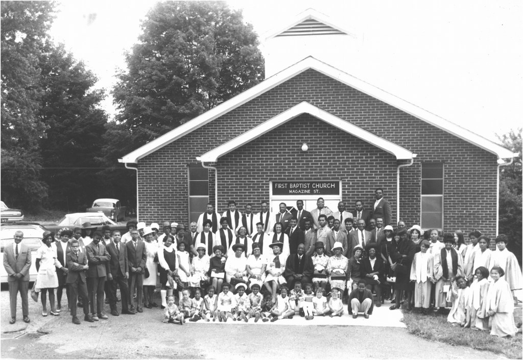 Black and white photo of a church congregation standing outside of their church. Signage indicates this is the First Baptist Church on Magazine St. The congregation is dressed in Sunday best attire and choir robes. Individuals of all ages standing, kneeling, seated in chairs, and seated on the ground smiling at the camera.