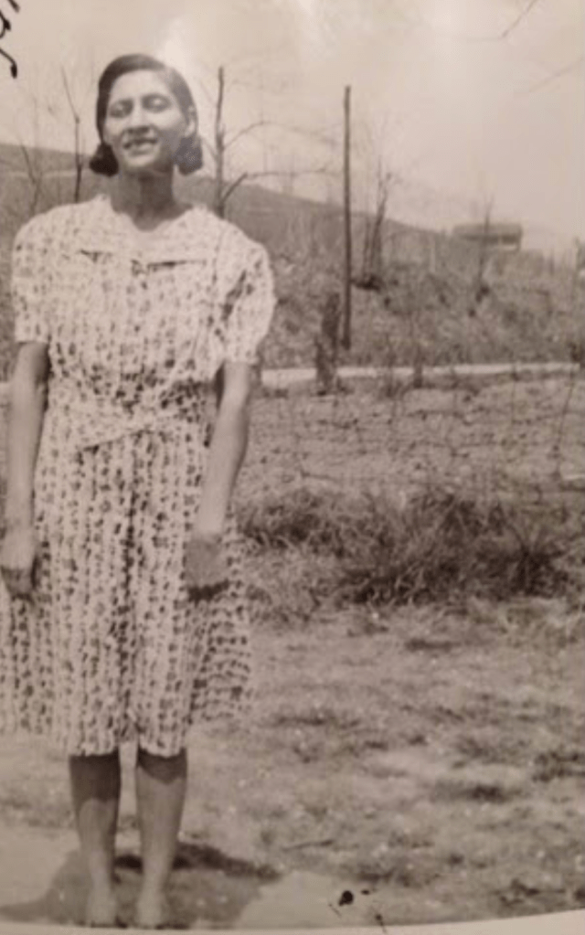 Black and white photo of a woman with short straight hair and side part standing in a field. She is wearing a white printed dress with a collar.