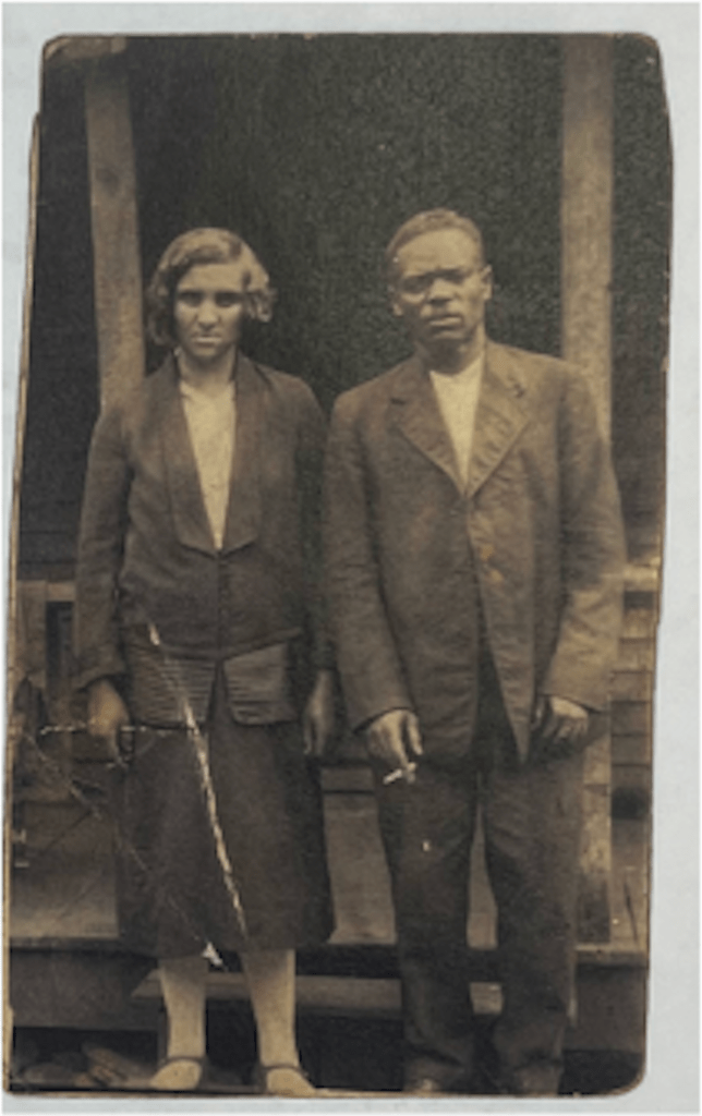 Black and white photograph of a couple standing outside of a wooden structure. The woman with short hair on the left is wearing a coat and skirt with stockings squinting. The man on the right is wearing a suit jacket and slacks with a cigarette between his fingers with a furrowed expression. 