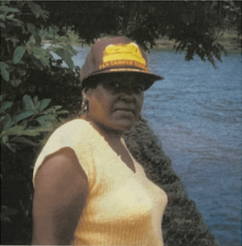 Photo of a woman standing on the bank of a body of water, facing the water wearing a yellow shirt and brown and yellow hat looking over her right shoulder to face the person taking the photo 