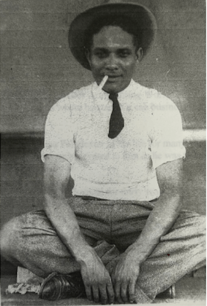 Black and white portrait of a man seated cross-legged with his hands over his hands draped over his ankles. He is waring a hat, ties, vest, button down shirt with the sleeves rolled to his upper arm, and relaxed pants with combat boots. He is smiling with a cigarette handing out of the left side of his mouth. 