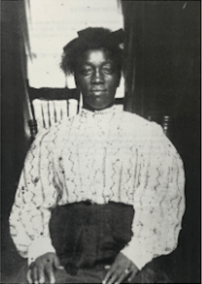 Black and white portrait of a woman sitting in the foreground wearing a patterned white blouse and a dark skirt. Her hans are placed in her lap and she is gazing directly ahead. In the background sits a wooden chair in front of a window with dark curtains.  