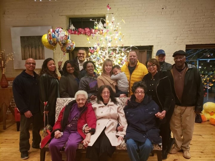 Picture of 14 family members. Front row has three older women sitting on a couch. The middle row has 6 adult s standing and one of them is holding a young child. The back row has four adults standing staggered. There is a Christmas tree in the background.