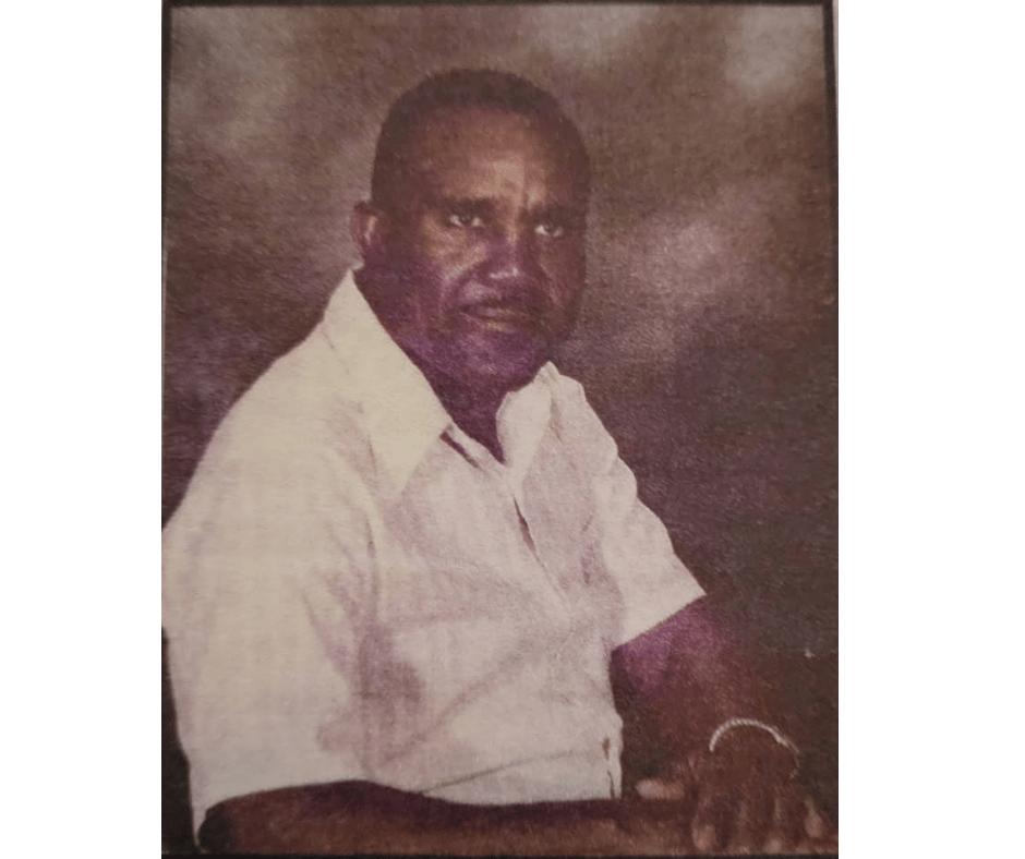 Portrait of a man sitting at an angle to the camera wearing a white dress shirt and silver watch. He is posed with his arms crossed on hand over the other, and he has a neutral expression. 
