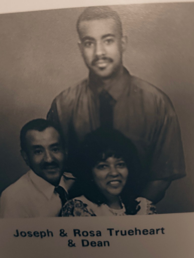 Photo of three individuals captioned "Joseph & Rosa Trueheart & Dean" Joseph and Rosa are sitting wearing nice clothing and Dean is standing behind them also with a shirt and tie. They are all smiling. 