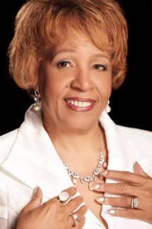 Colorized portrait of a woman wearing a white blazer and extravagant jewelry. She is posing with her arms rested on her upper chest. 