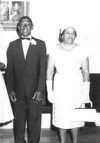 Black and white photograph of a man and a woman. The man is on the left dressed in a black suit with a radiant smile. The woman is on the right dressed in a white knee-length dress with white gloves, white purse, and pearls. The woman is gazing to her left.