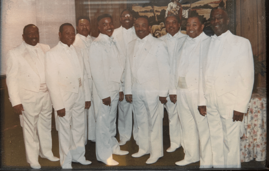 Nine men standing staggered in a v shape. All wearing white tuxedos and white dress shoes. 