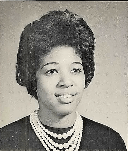 Black and white yearbook portrait of a young woman. She has intricately styled hair and is wearing four strings of pearls that descend in size. She is smiling softly and gazing to her left.