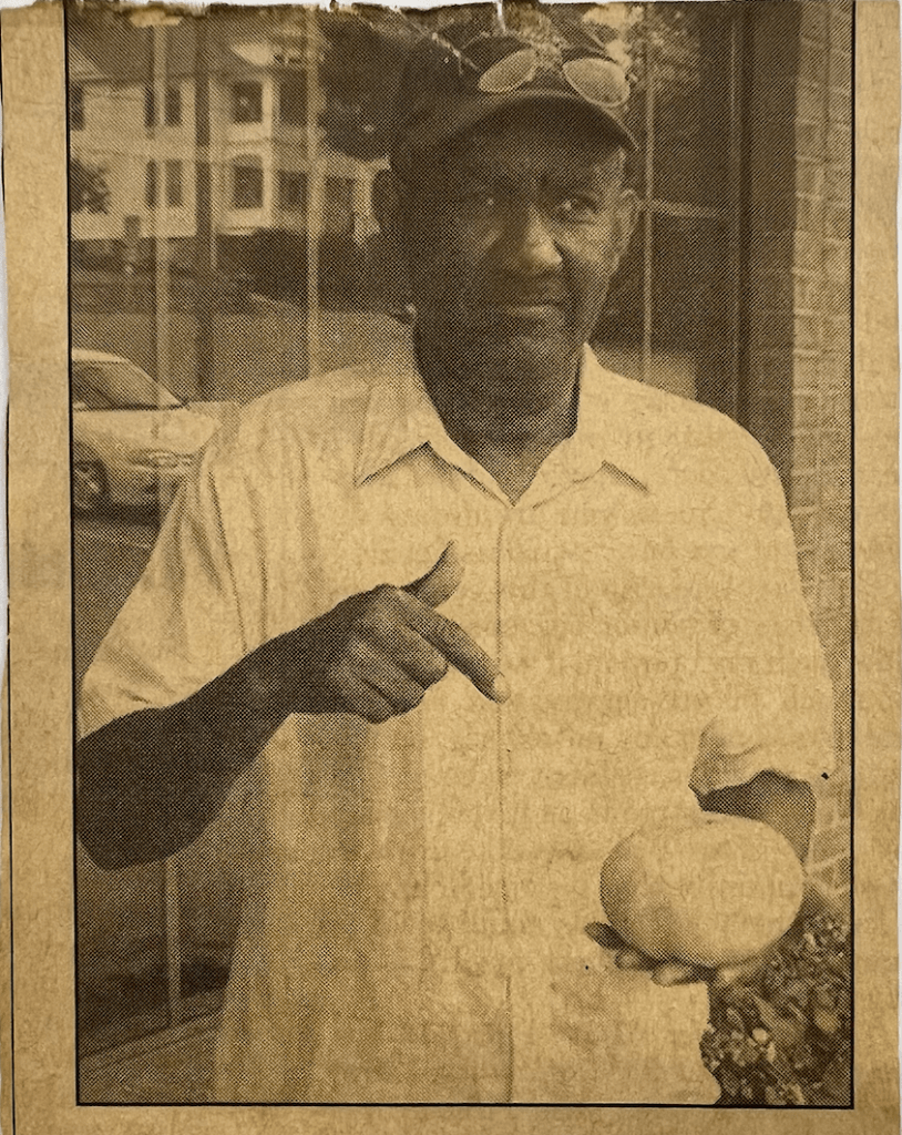 Photo of a newspaper clipping. A man is standing wearing a hat with sunglasses draped atop the hat and a white button-down shirt. He is holding an oversized tomato in his left and pointing to the tomato with his right hand. 
