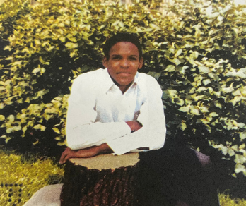 Image of Tony Conner in a white button down, sitting in the grass and leaning on tree stump.