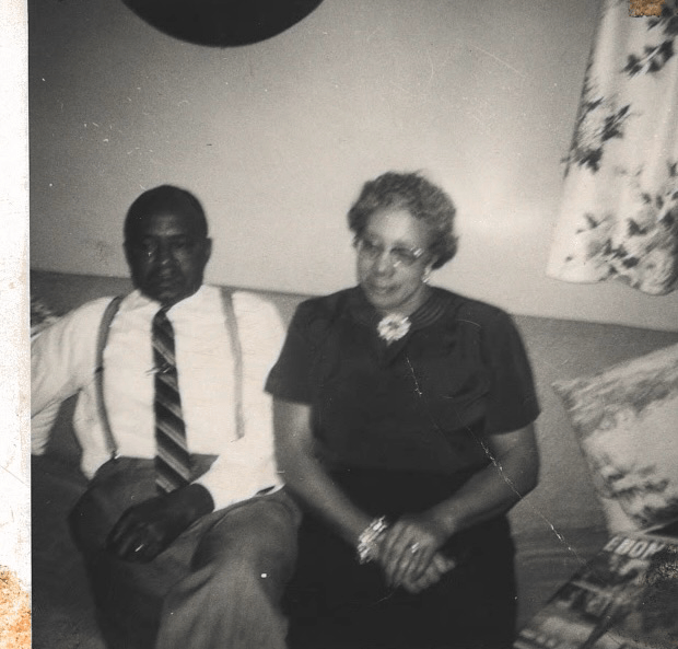 A man and woman sit on couch together dressed in formal clothes. The man wears suspenders and a tie and the woman wears a black dress. Both gaze left