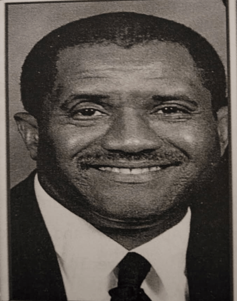 Portrait of a man wearing a suit jacket, white dress shirt, and black tie smiling at the camera.