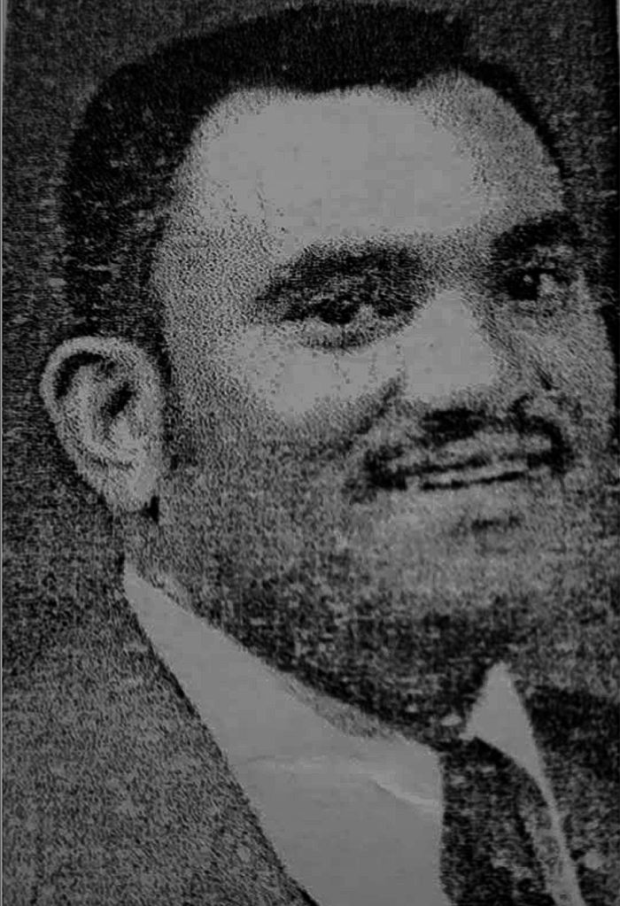 Black and white photo of a man smiling for a portait wearing a suit and tie. He is posed at an angle looking over his right shoulder. 